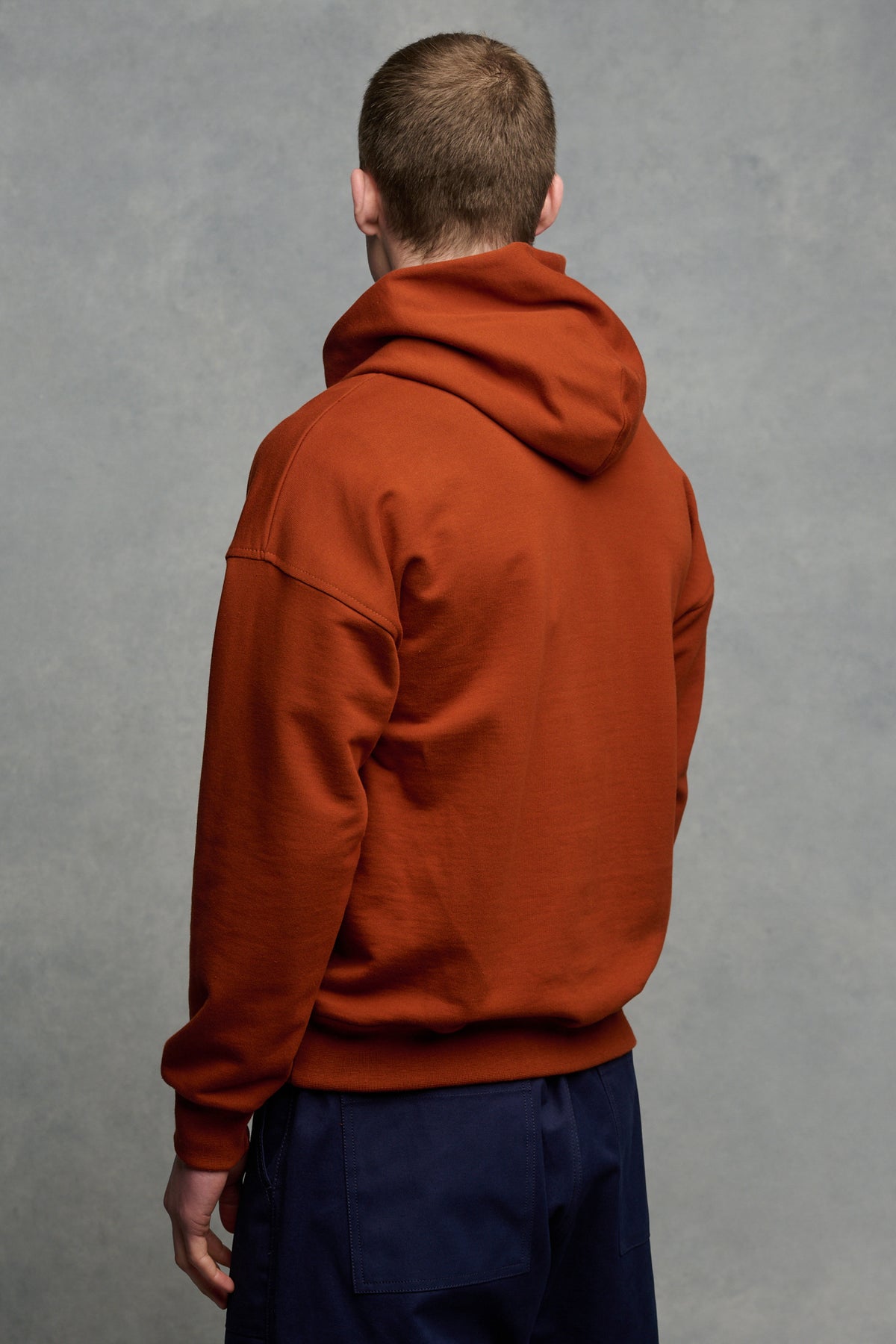 The back of brunet male wearing hooded sweatshirt in cinnamon paired with navy cameraman pant