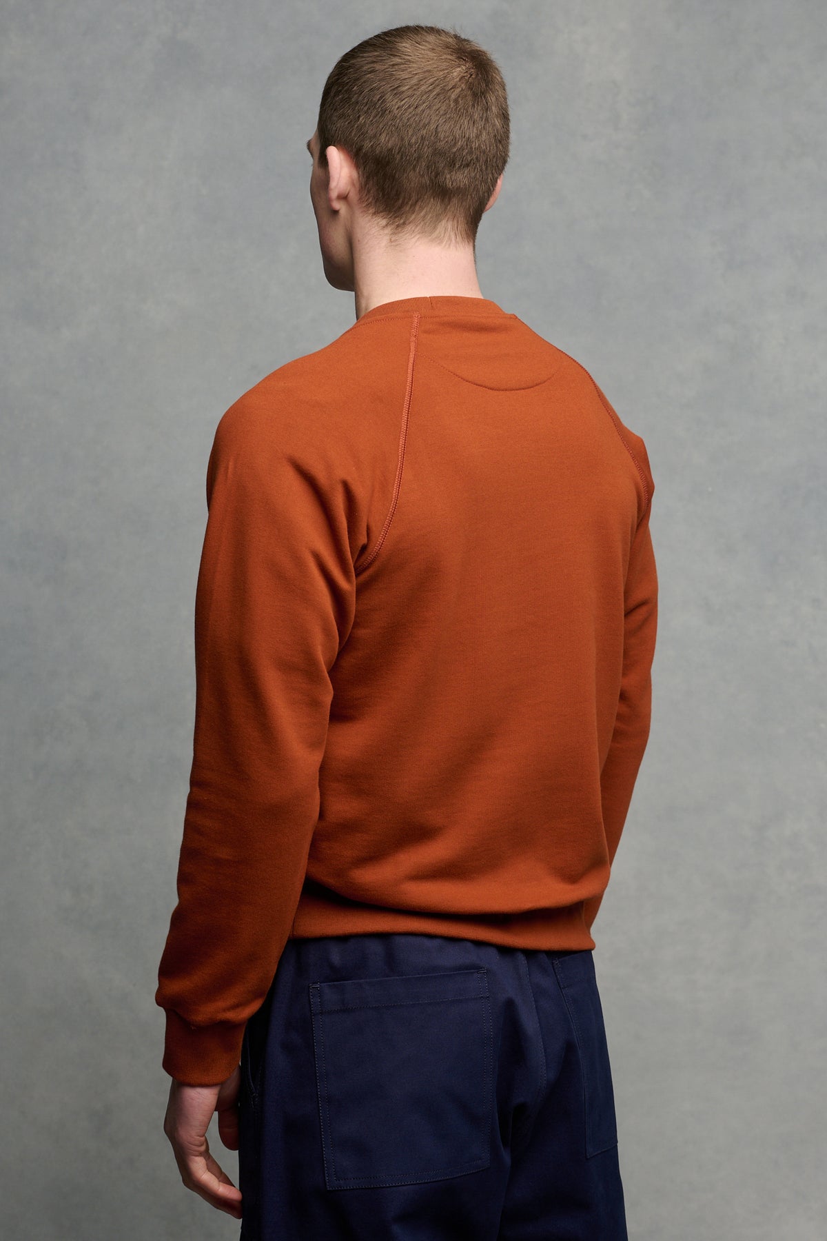 The back of male wearing men's raglan sweatshirt in cinnamon paired with navy cameraman pant