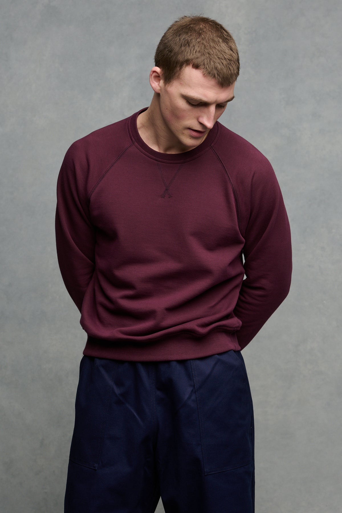 Thigh up image of male looking down to ground with hands behind his back wearing raglan sweatshirt in plum