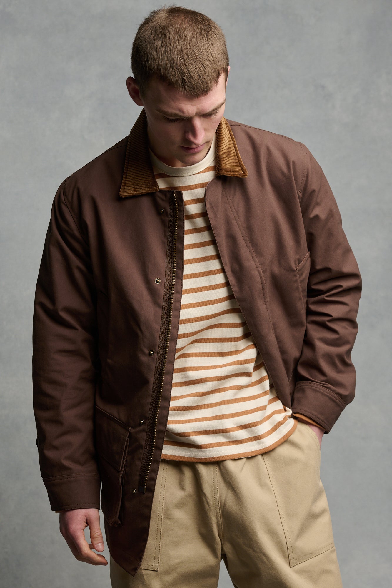 Thigh up image of male wearing men's car coat in brown unzipped over Breton in ecru cinnamon paired with cameraman pant in putty