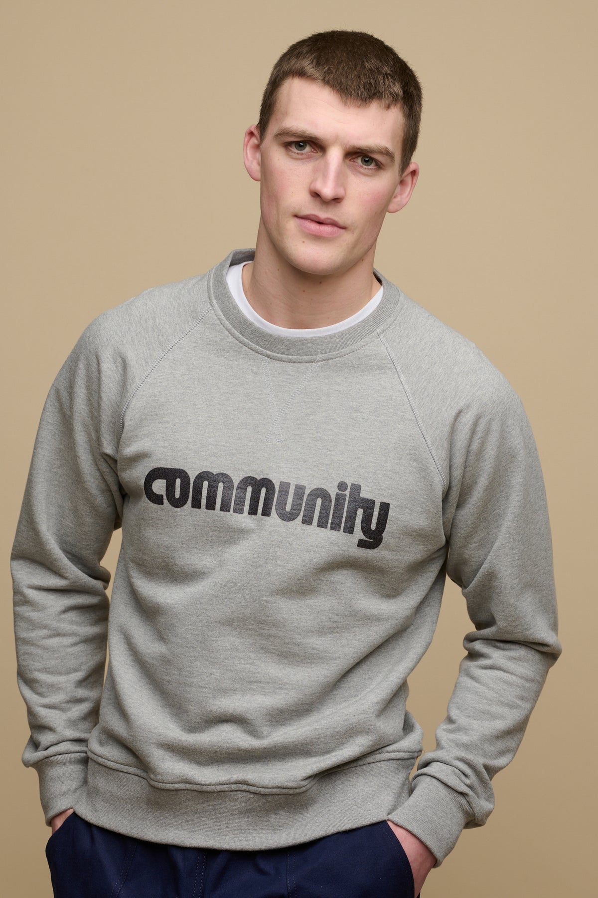 
            Front of brunet male wearing logo raglan sweatshirt in grey marl with Community logo across the chest, crew neck t shirt in white visible worn underneath.