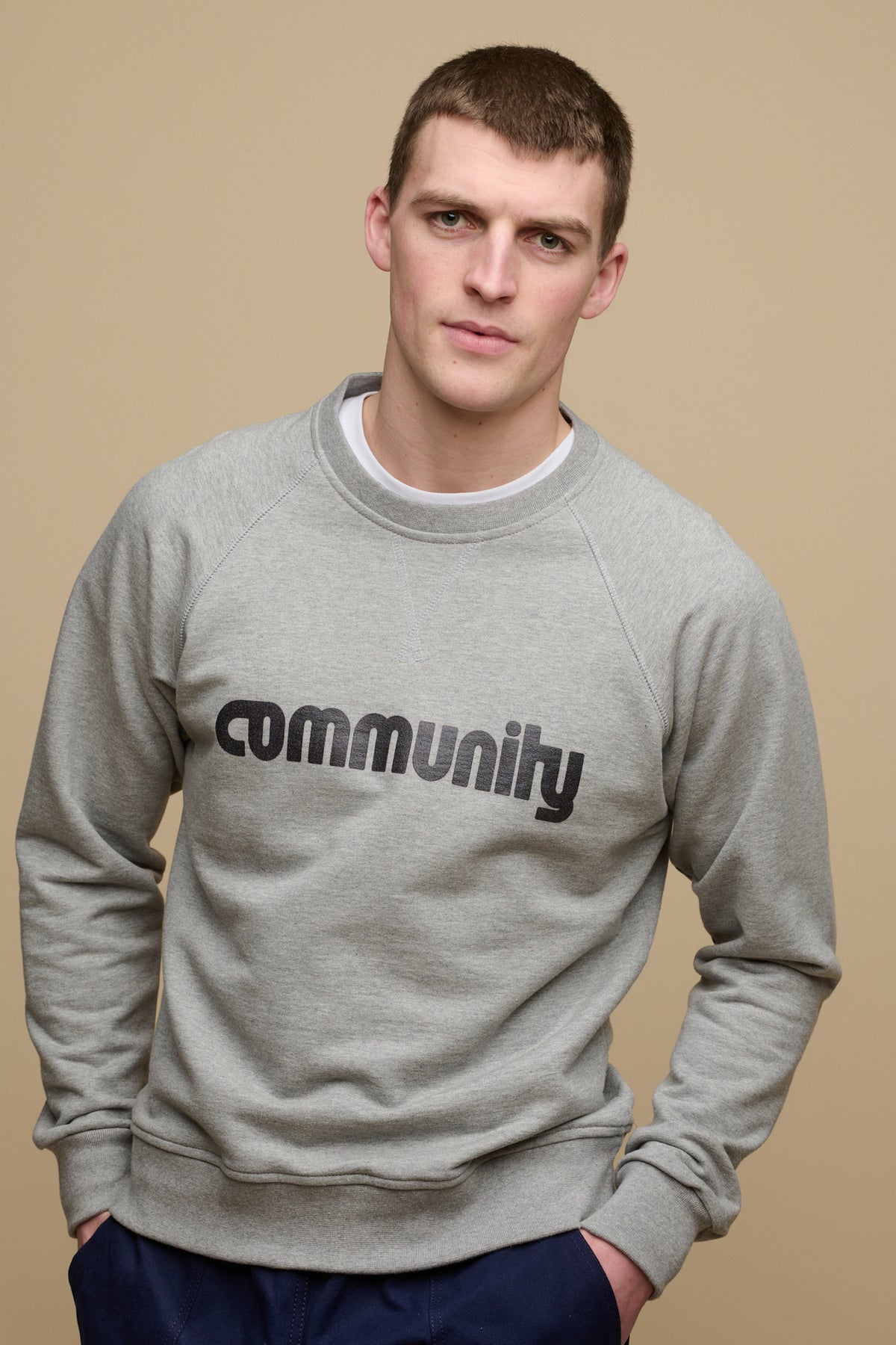 Front of brunet male wearing logo raglan sweatshirt in grey marl with Community logo across the chest, crew neck t shirt in white visible worn underneath.