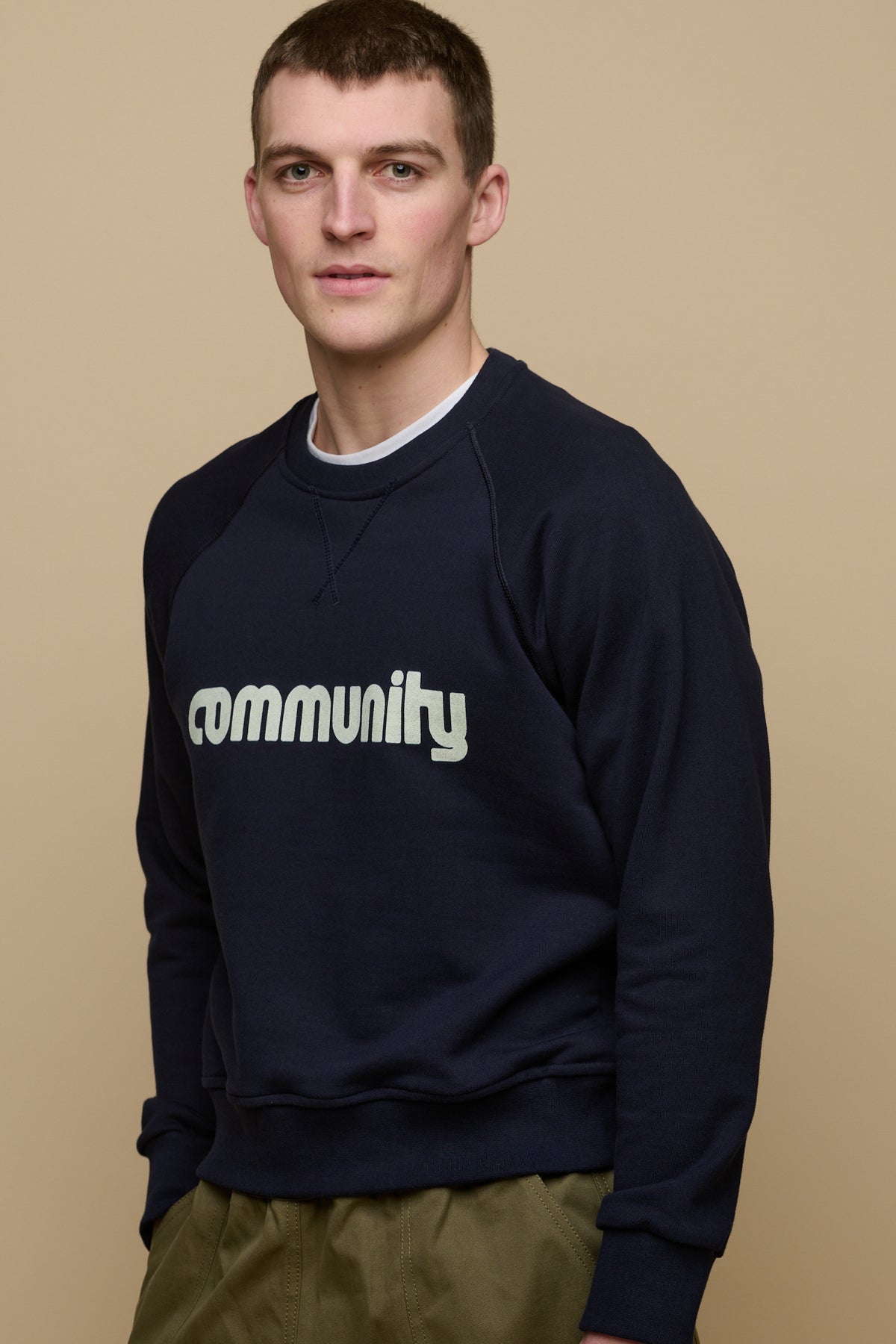 Thigh up front of brunet male wearing logo raglan sweatshirt in navy worn over white crew neck t shirt paired with olive cameraman pant