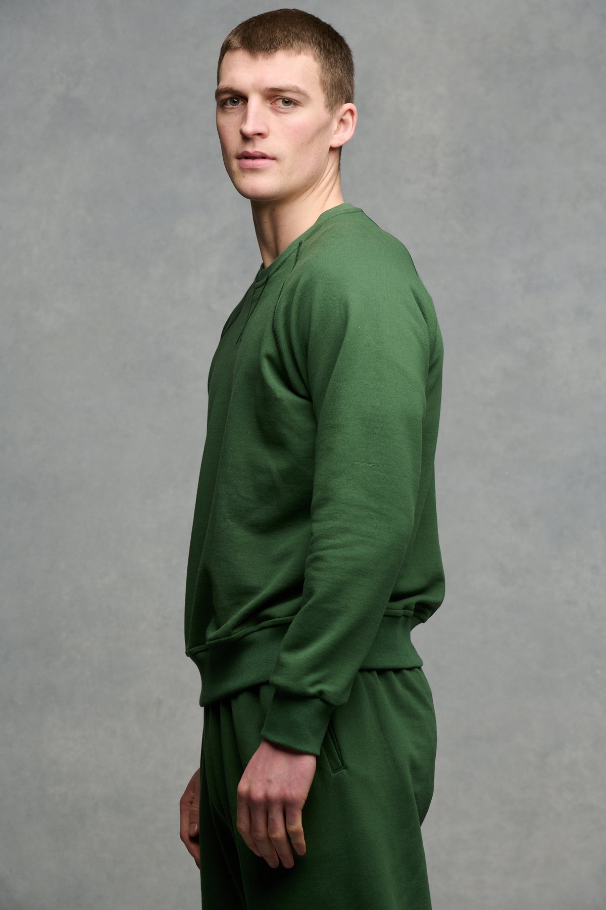 The side of male from the thigh up wearing raglan sweatshirt in bottle green paired with matching sweatpant in bottle green