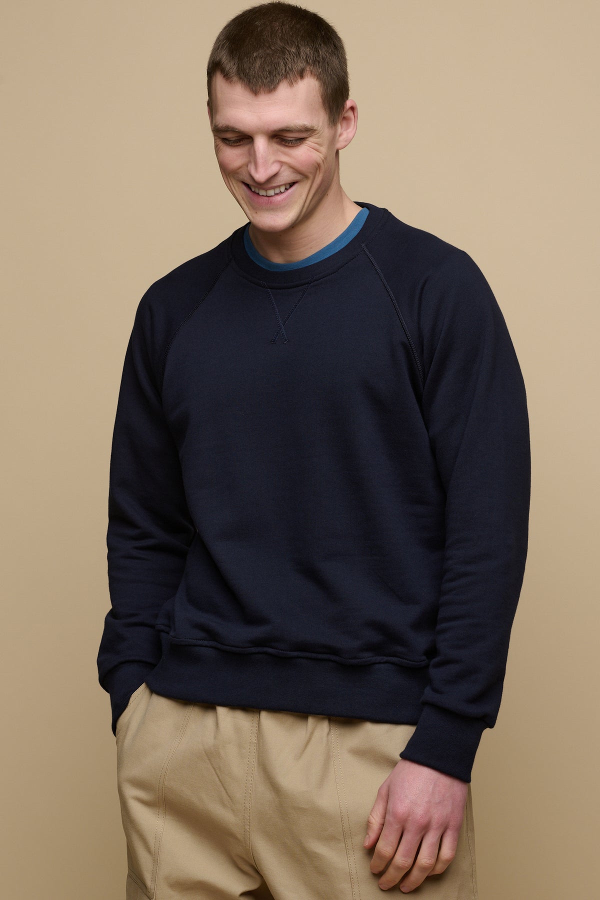 Thigh up image of smiley brunet male wearing raglan sweatshirt in navy worn over crew neck t shirt in raf blue paired with cameraman pant in putty