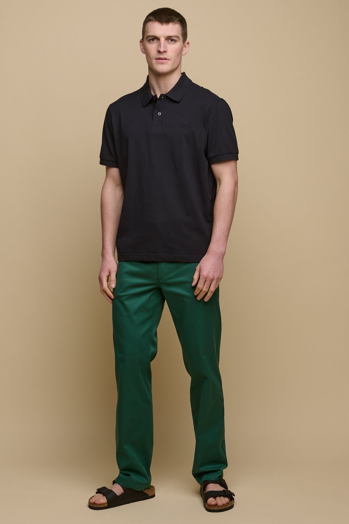 Full body image of brunet male wearing men's slim stretch chino in bottle green paired with short sleeve polo shirt in navy and sandals