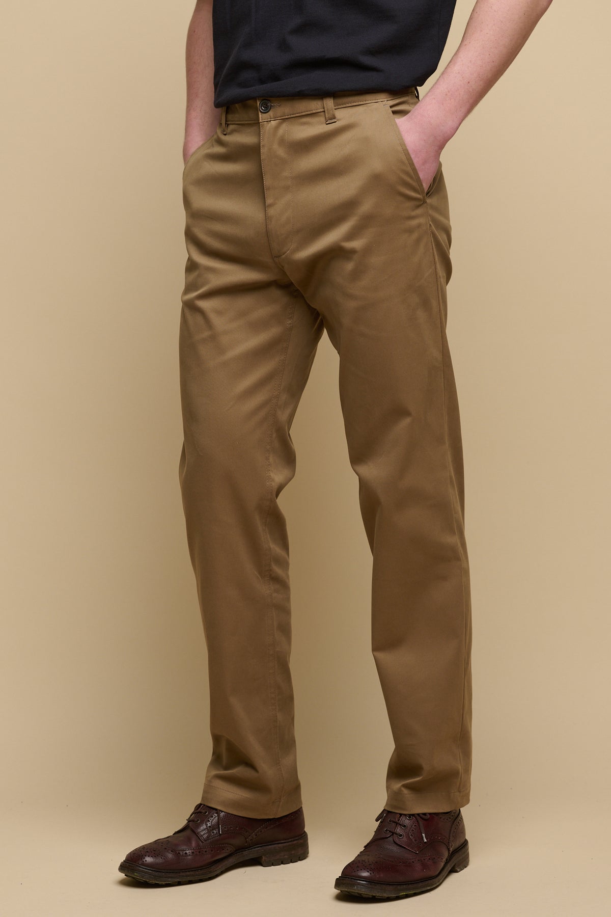 Chest down of male wearing slim stretch chino in khaki with hands in front pockets worn with brown leather shoes