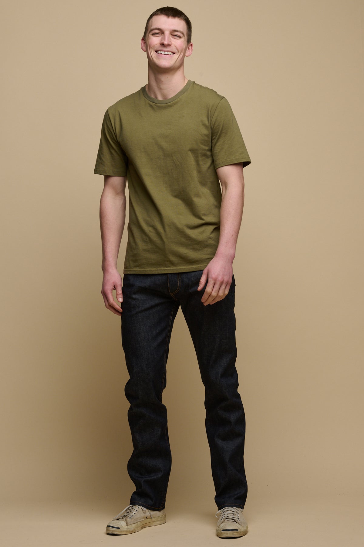 
            Full body shot of smiley male wearing straight cut selvedge jeans in raw denim paired with short sleeve t-shirt in olive and trainers
