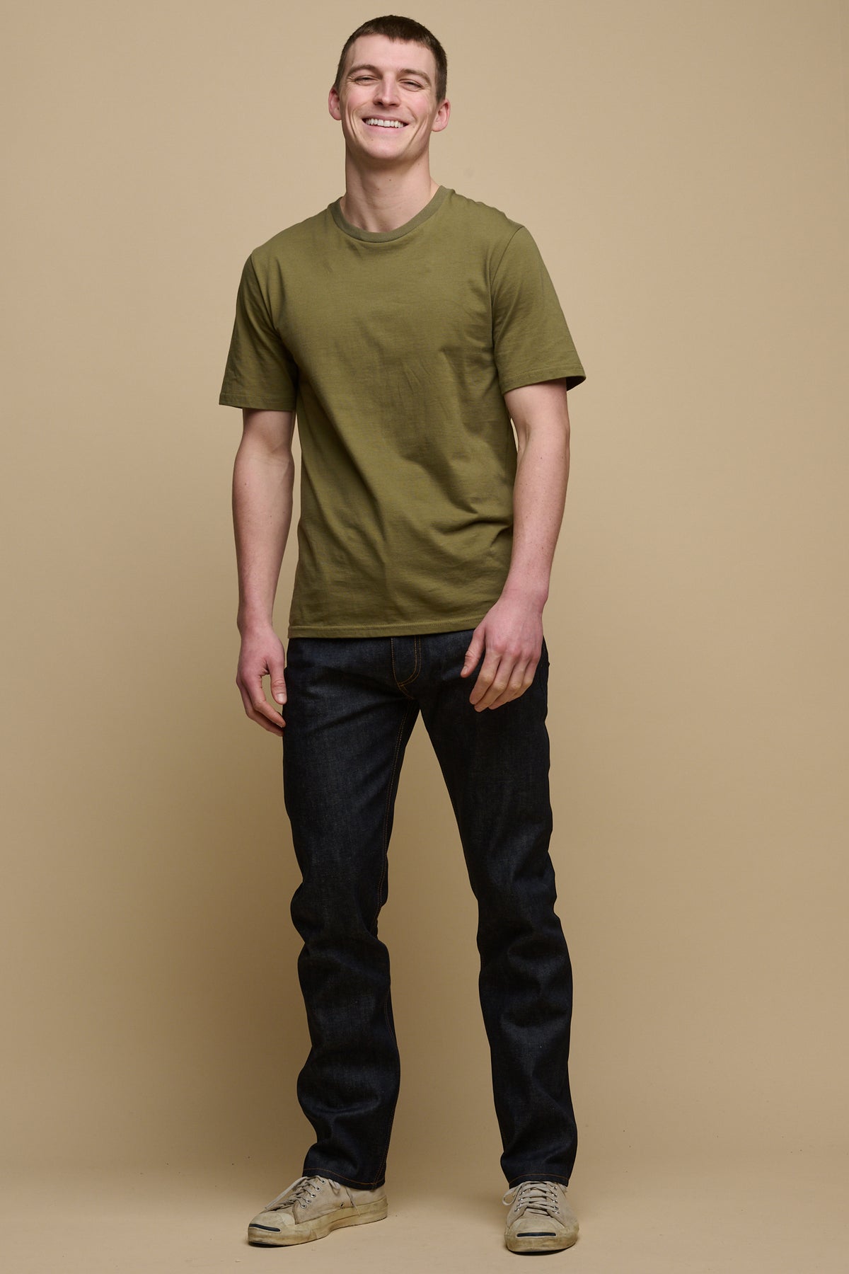 Full body shot of smiley male wearing straight cut selvedge jeans in raw denim paired with short sleeve t-shirt in olive and trainers