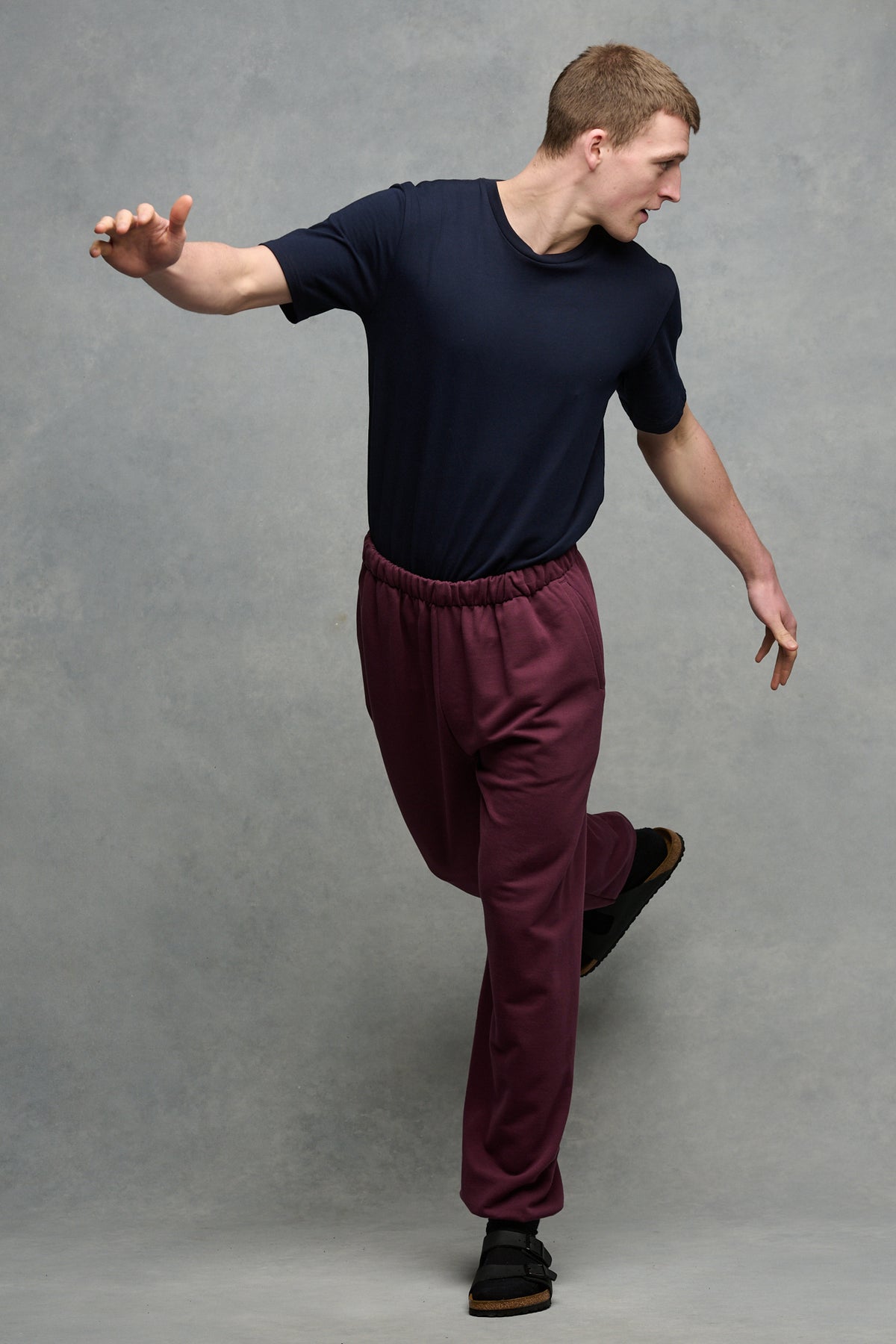 Full body image of male balancing on one leg wearing sweatpants in plum with navy crew neck t-shirt tucked in