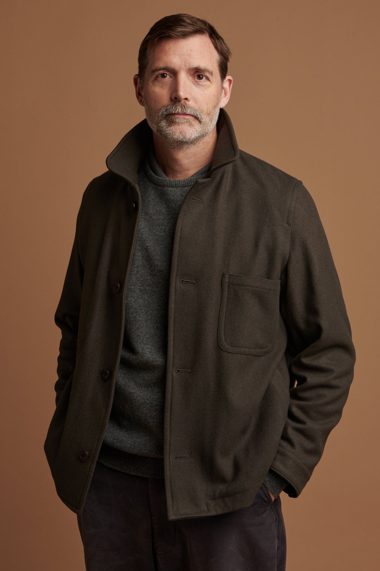 Thigh up of Patrick wearing Arthur wool chore jacket in the colour drab. Button down fastening with three external pockets. Worn over lambswool saddle shoulder crew neck jumper in grey