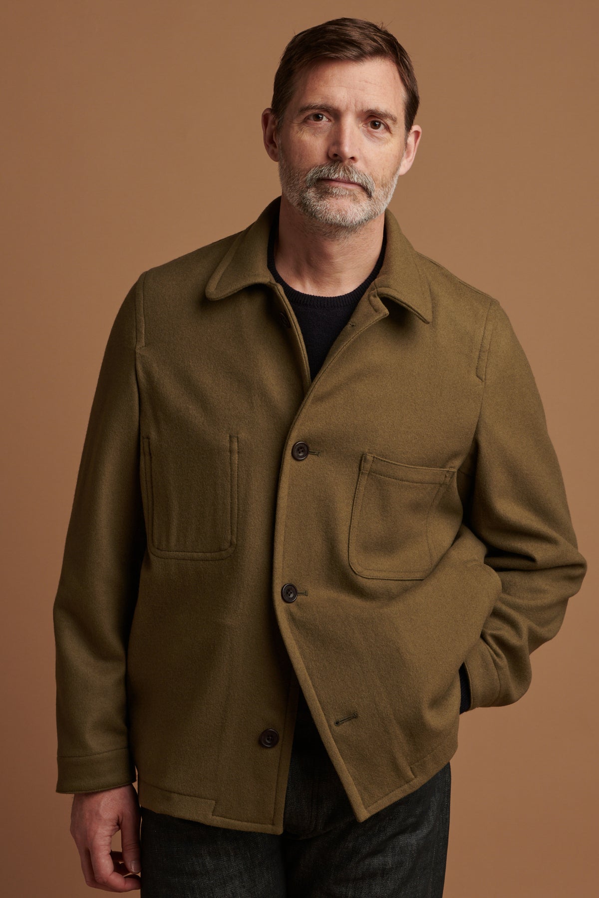 Patrick Grant facing the camera wearing Arthur wool chore jacket in olive with two out of four buttons fastened. Patrick wears the jacket over lambswool saddle shoulder crew neck in navy