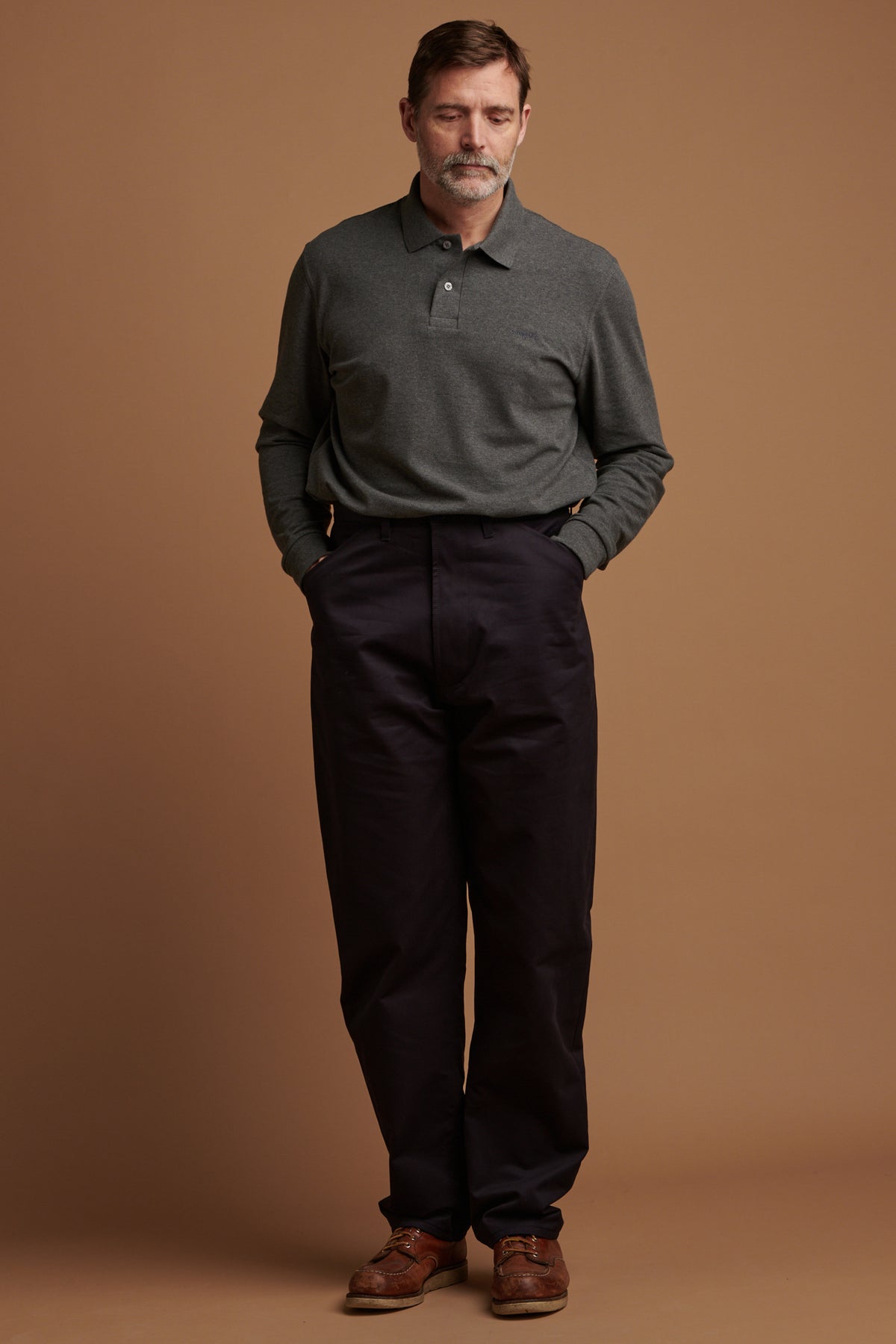 
            Patrick wearing cotton chore trousers with his hands in the front pockets paired with long sleeve polo shirt in grey