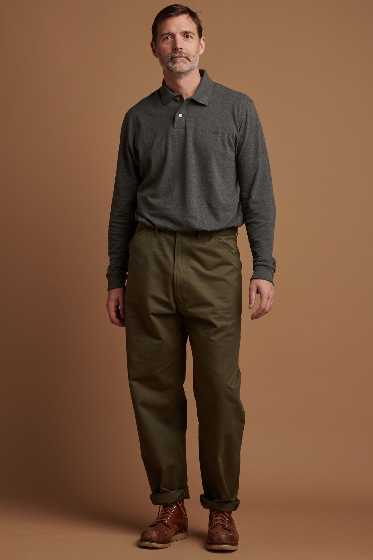 
            The front of Patrick looking into the camera wearing cotton chore trousers in olive with a rolled hem, paired with long sleeve polo shirt in grey tucked into trousers