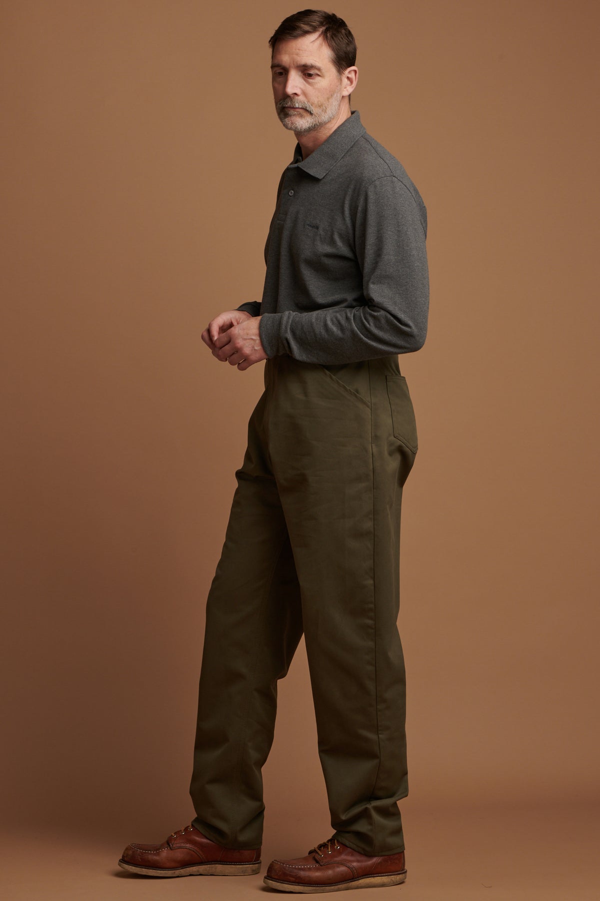 
            Patrick Grant facing to the left wearing cotton chore trousers in olive paired with Community Clothing long sleeve polo shirt in grey and brown leather shoes