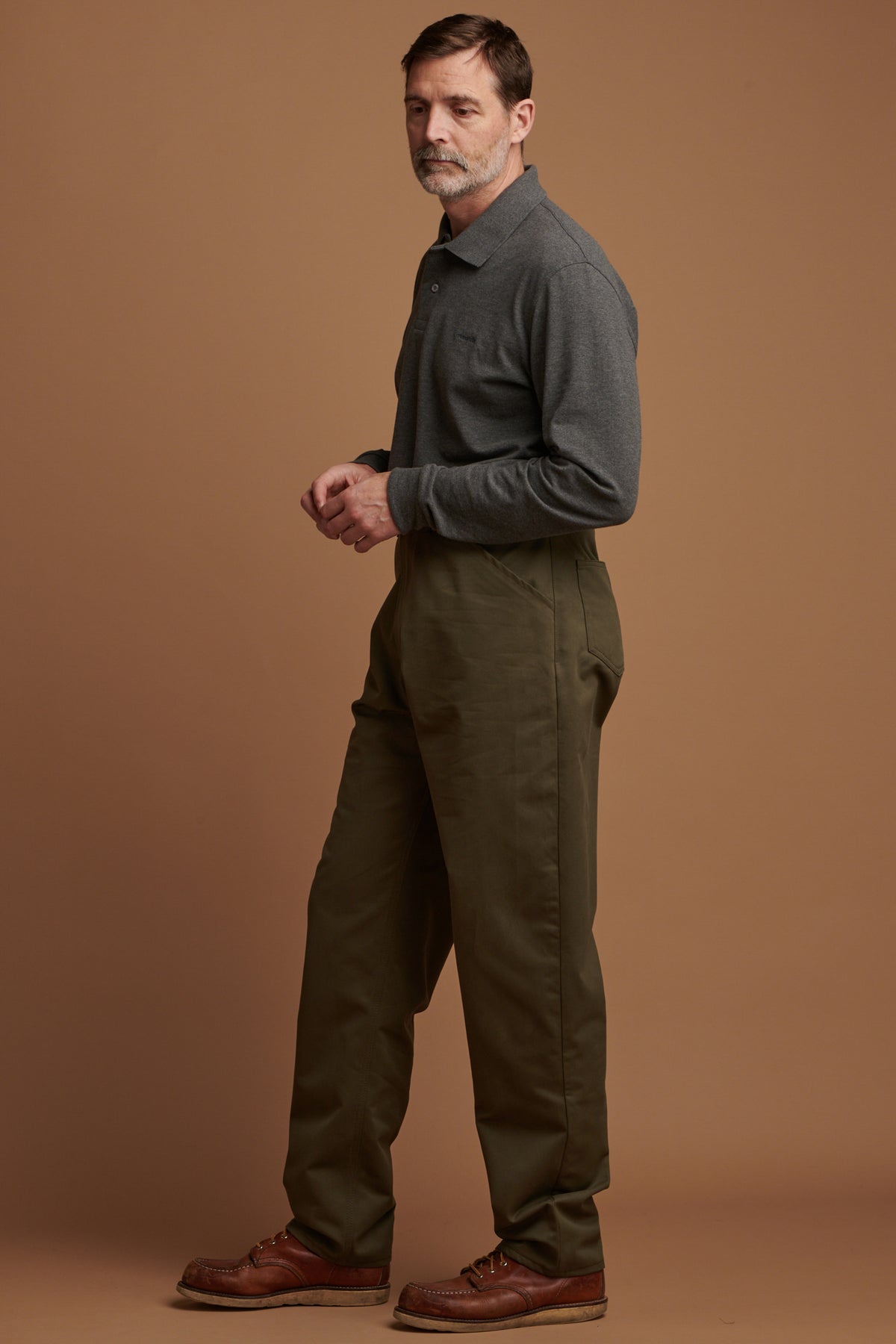 Patrick Grant facing to the left wearing cotton chore trousers in olive paired with Community Clothing long sleeve polo shirt in grey and brown leather shoes