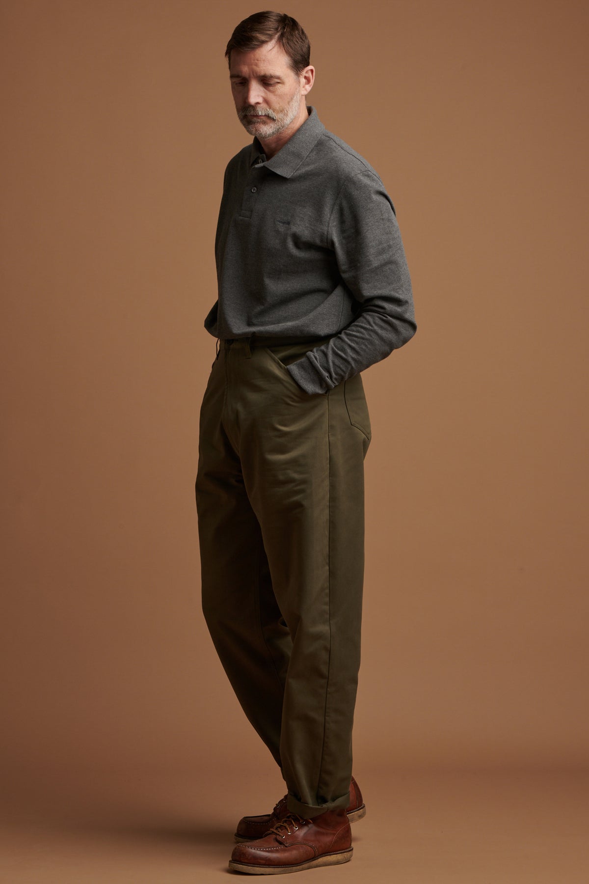 Patrick Grant facing to the left with his hand in the front pocket of cotton chore trousers in olive paired with Community Clothing long sleeve polo shirt in grey and brown leather shoes