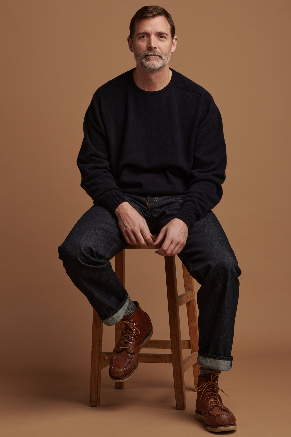 
            Patrick Grant sat on stool wearing loose fit button fly Arran jean in indigo paired with lambswool saddle shoulder crew neck in navy