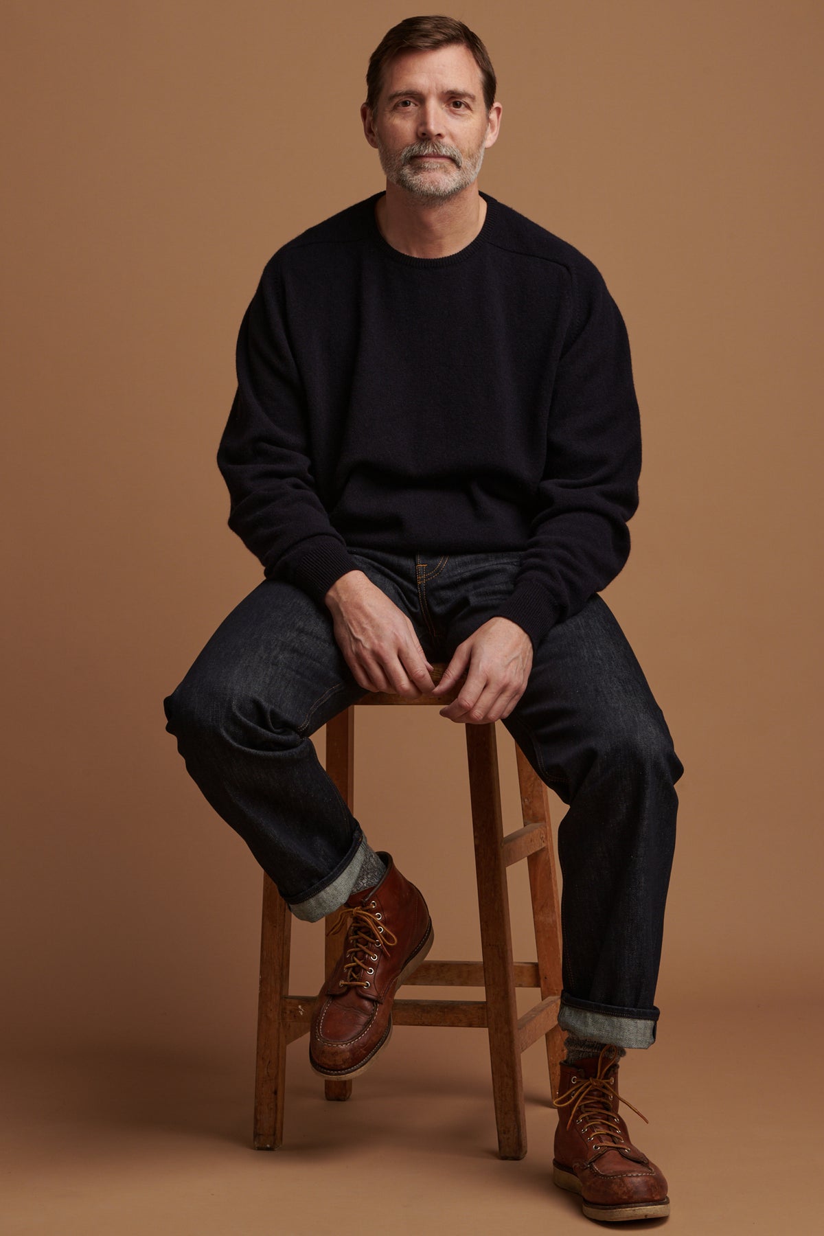 Patrick Grant sat on stool wearing loose fit button fly Arran jean in indigo paired with lambswool saddle shoulder crew neck in navy