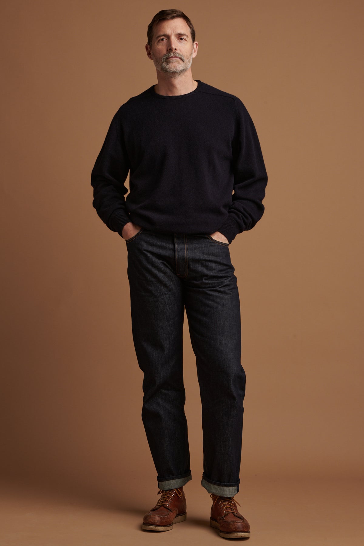 The front of Patrick wearing loose fit button fly Arran jean in indigo with his hands in the front pockets and the hem rolled up, paired with lambswool saddle shoulder crew neck in navy.