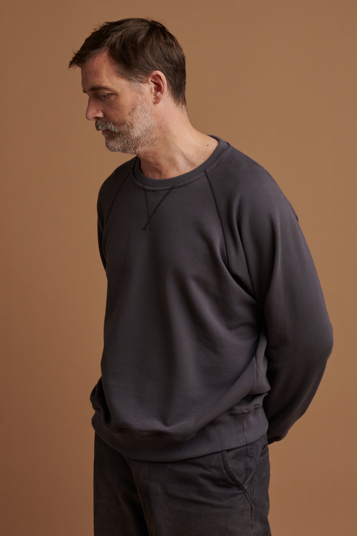 Patrick Grant wearing organic cotton raglan sweatshirt in slate grey looking towards the ground with his arms behind his back