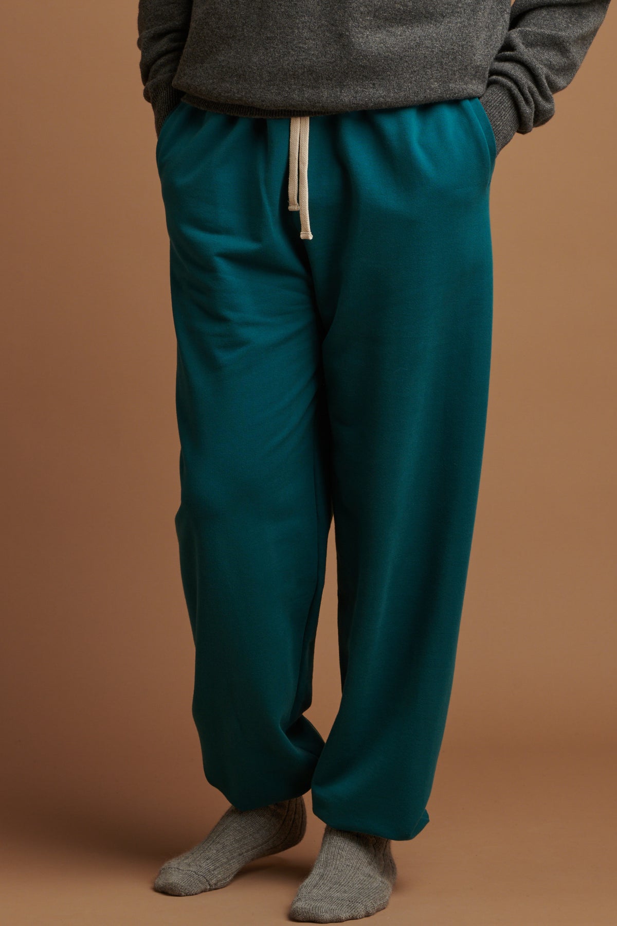 Waist down of the front of male wearing men's sweatpants in teal, with hands in the front pockets and ecru drawcord on show, paired with alpaca bed socks