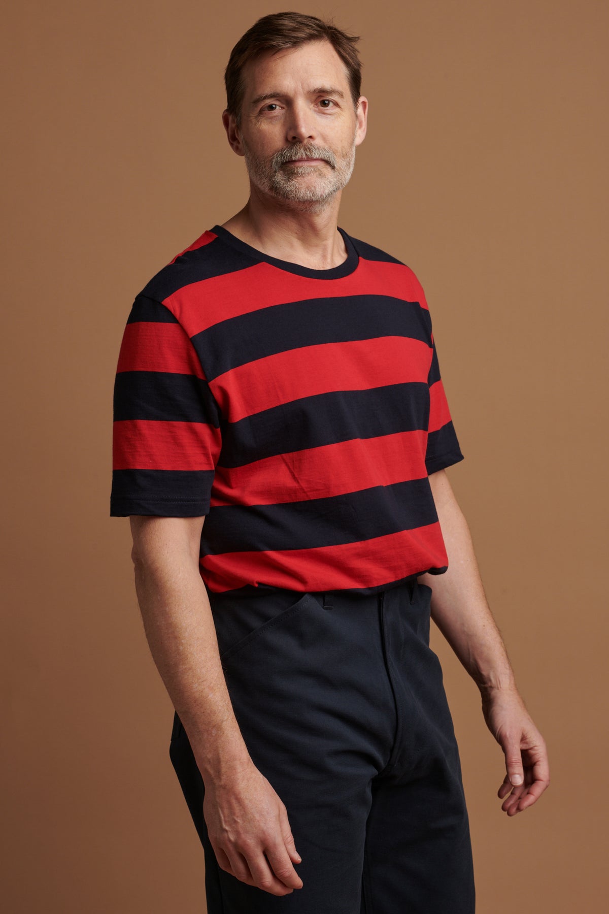 
            Thigh up of Patrick Grant wearing Community Clothing wide stripe short sleeve t shirt in navy and red tucked into navy chore trousers 