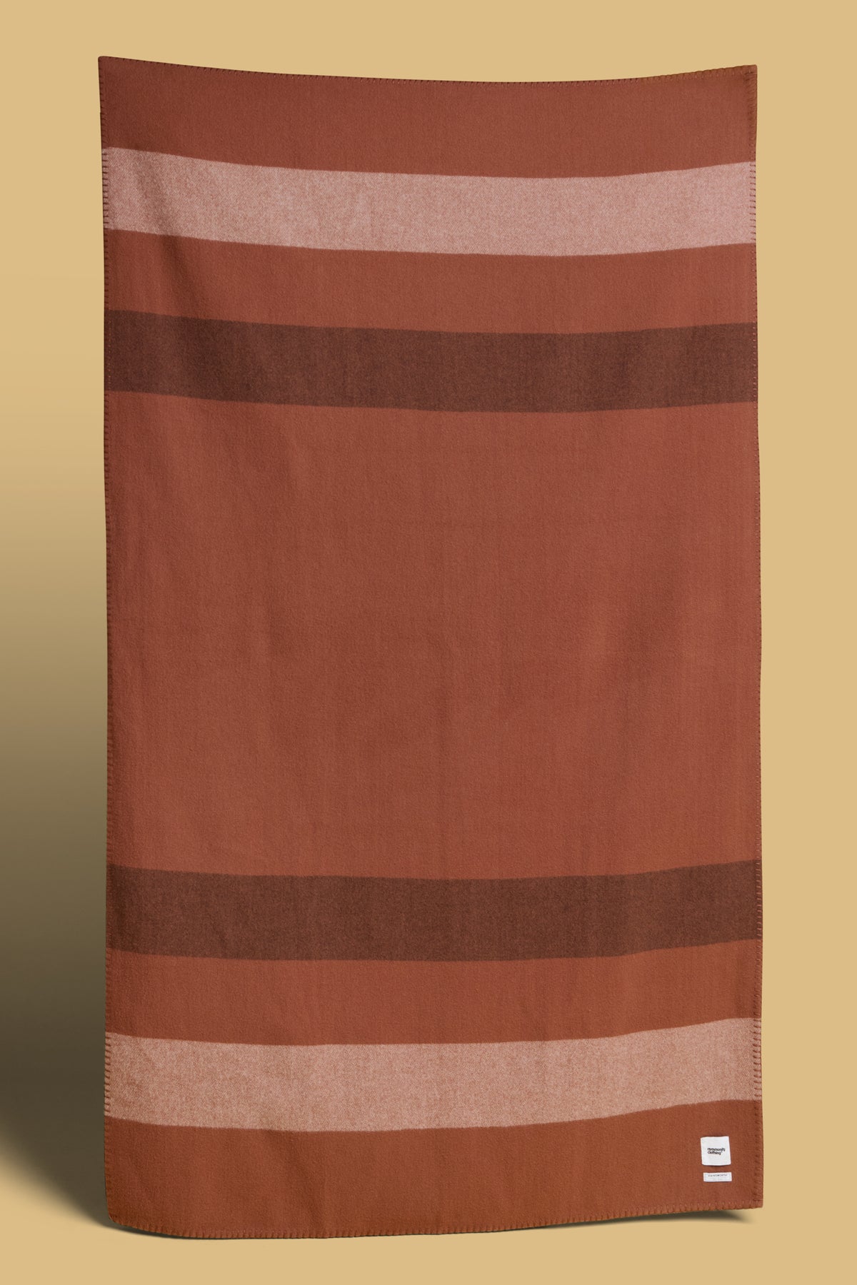 
            Blanket in tan with dark brown and ecru stripes, Community Clothing logo in bottom right hand corner of the blanket.