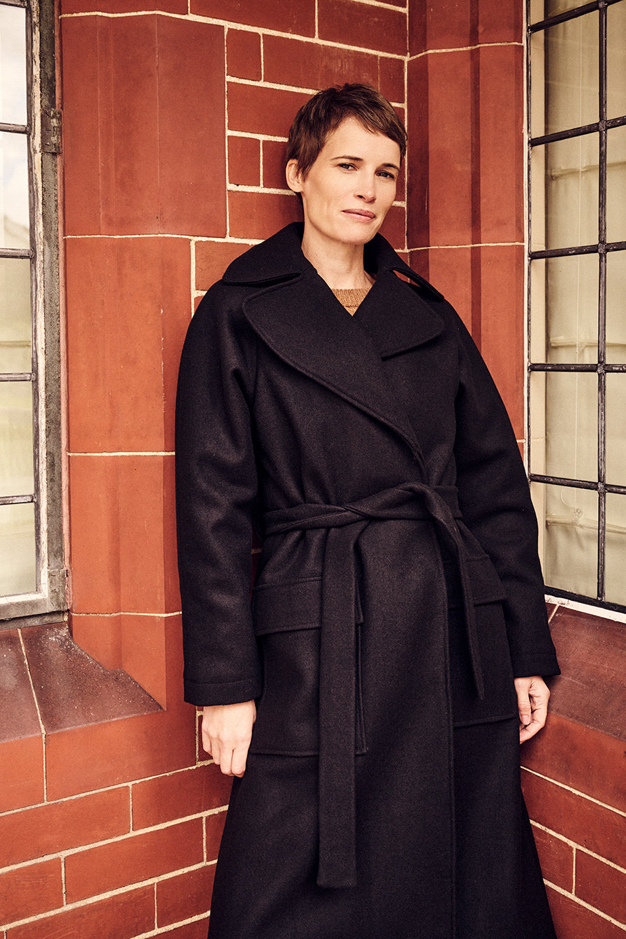 Woman wearing a black coat standing against a red brick wall.